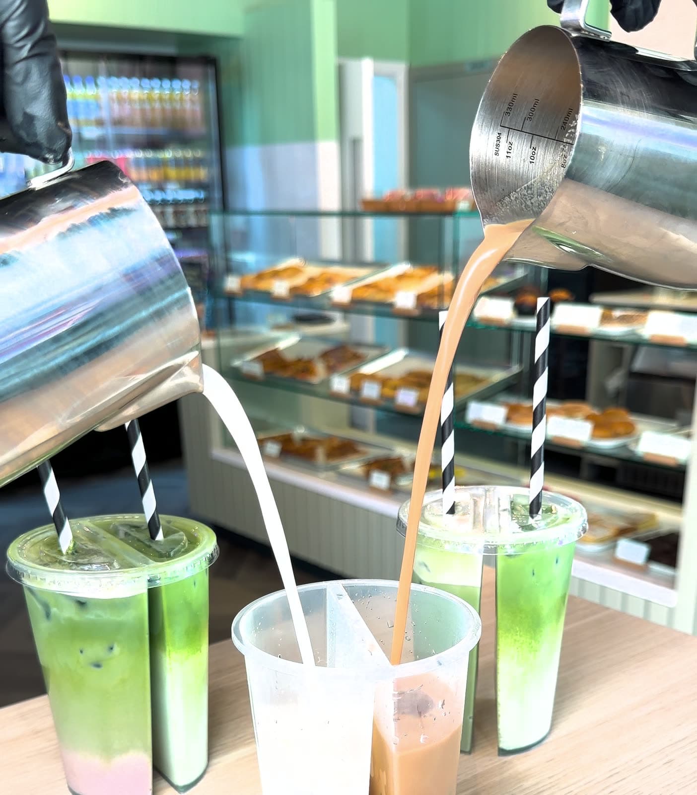 Two jugs pouring matcha and coffee into a split cup at the counter